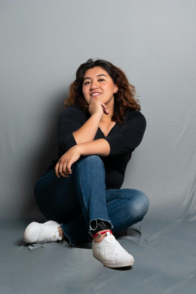 A cheerful woman posing casually in jeans and sneakers against a plain backdrop.