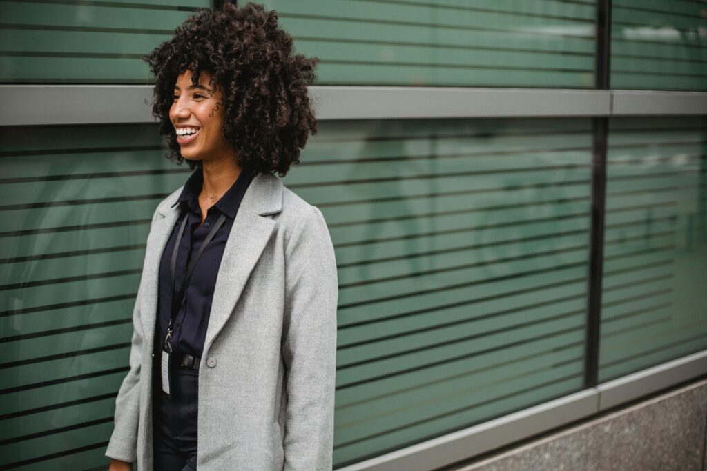 A woman with afro hair smiles confidently, wearing a stylish coat against an urban backdrop.