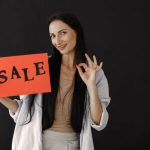Brunette woman gesturing okay while holding a red sale sign on black background, perfect for consumerism and shopping themes.